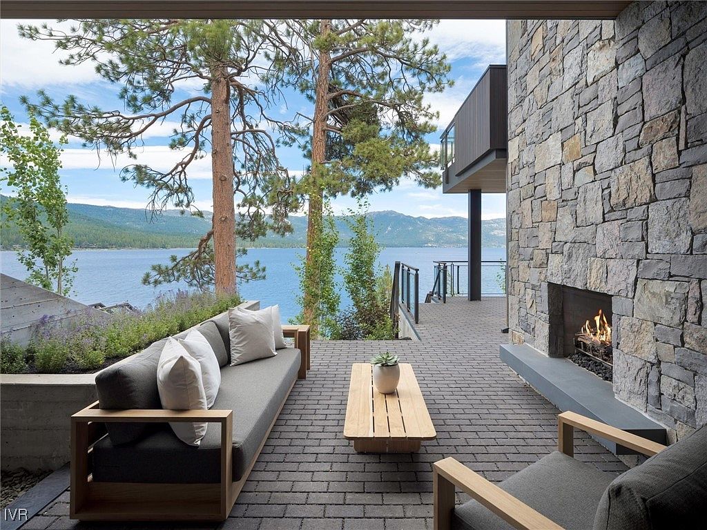 This image showcases a luxurious outdoor patio area with a stunning lake view. The patio features a stone fireplace, comfortable seating, and a wooden coffee table, creating an inviting space for relaxation and entertainment. The design incorporates natural elements, blending seamlessly with the surrounding landscape.