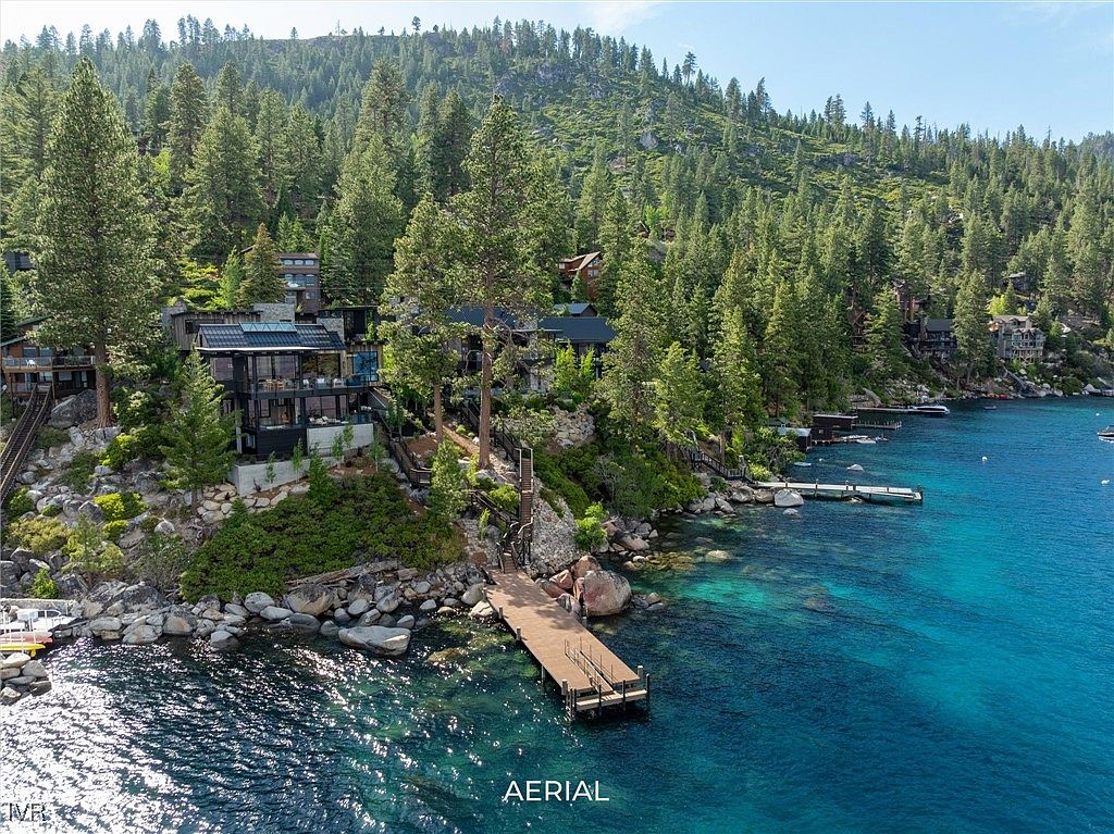 This aerial view showcases a luxurious waterfront property nestled among lush greenery. The modern architecture of the home blends seamlessly with the natural landscape, featuring multiple levels and outdoor living spaces. A private dock extends into the clear, turquoise water, offering direct access to the lake.