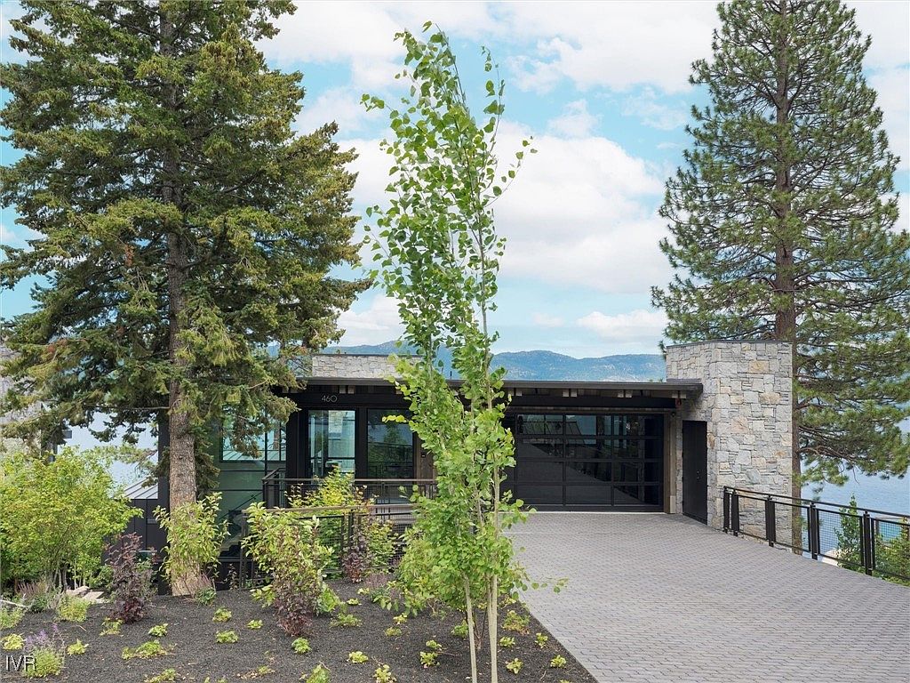 This is a front exterior view of a modern home with a stone facade and a black framed glass garage door. The property features mature trees and landscaping, with a paved driveway leading to the garage. The home appears to be situated near a lake or body of water, with mountains visible in the background, suggesting a scenic and private location.