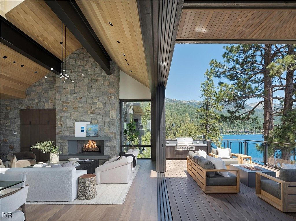 This image showcases a luxurious living room that seamlessly transitions to an outdoor deck. The interior features a stone fireplace, modern furnishings, and a wood-paneled ceiling, while the deck offers a built-in grill, comfortable seating around a fire pit, and stunning lake views. The open design creates an inviting space for both relaxation and entertainment.