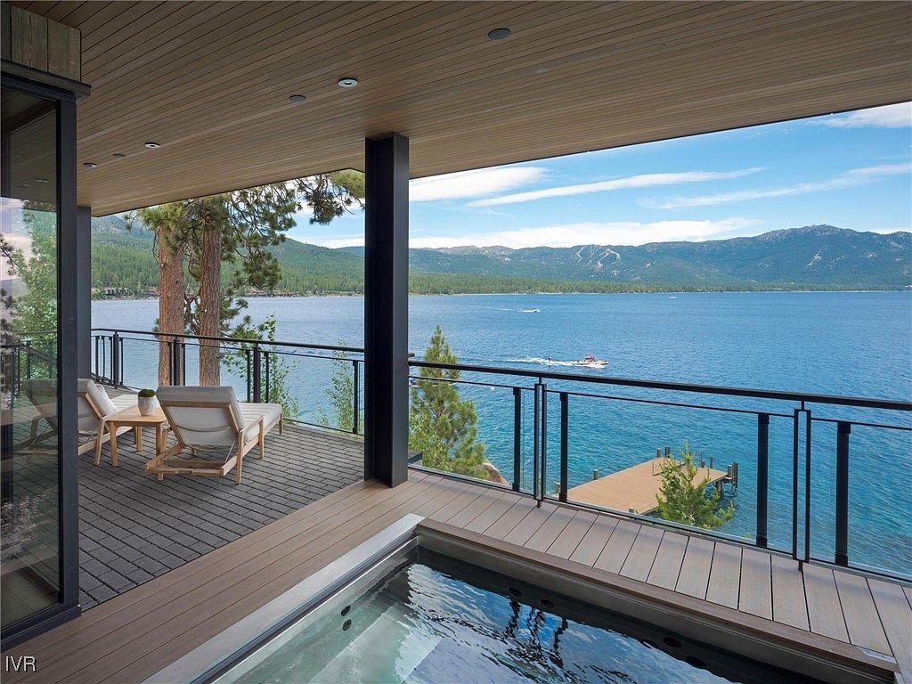 This image showcases a luxurious outdoor patio/deck area with a stunning lake view. The deck features modern lounge chairs, a hot tub, and a glass railing that provides an unobstructed view of the water and mountains in the distance. The space is designed for relaxation and entertainment, making it a desirable feature for potential buyers.