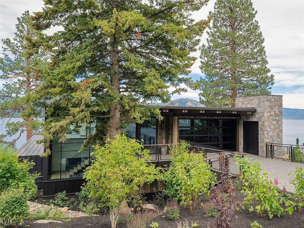 This is a front view of a modern home with a striking architectural design. The house features a combination of stone and dark wood siding, large glass windows, and a metal roof. A paved walkway leads to the entrance, surrounded by lush landscaping, creating an inviting and sophisticated curb appeal.