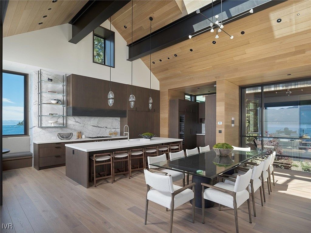 This is a spacious, modern kitchen and dining area featuring a large island with seating, sleek dark wood cabinetry, and a marble backsplash. The dining area includes a glass-topped table with seating for eight, and large windows offer a scenic view. The open floor plan and high ceilings with wood paneling create an airy and luxurious atmosphere.