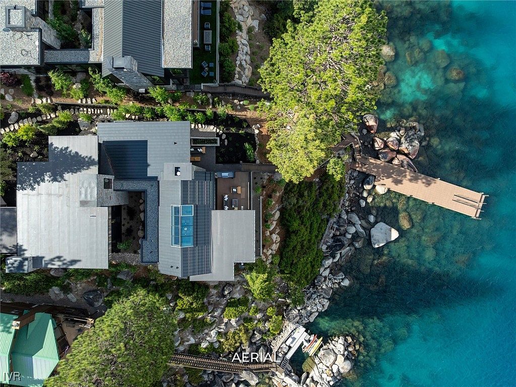 This aerial view showcases a luxurious waterfront property with a modern architectural design. The house features multiple interconnected structures with varying rooflines, a private dock extending into the clear turquoise water, and lush landscaping blending seamlessly with the natural rocky shoreline. The perspective offers a comprehensive view of the property's layout and its integration with the surrounding environment.