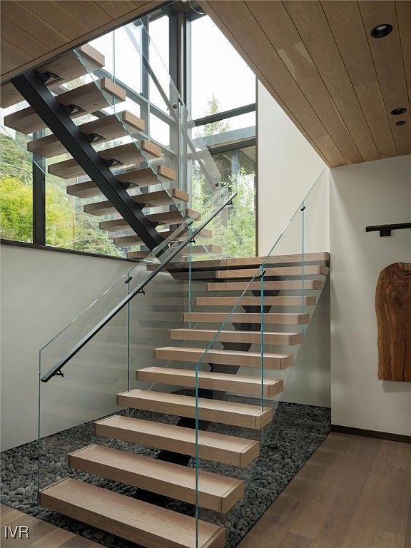 This interior shot showcases a modern staircase with floating wooden treads and glass railings, creating a minimalist and open design. The staircase is illuminated by natural light streaming through large windows, and the base of the stairs features a bed of dark pebbles, adding a unique textural element. The overall impression is one of contemporary elegance and architectural sophistication.