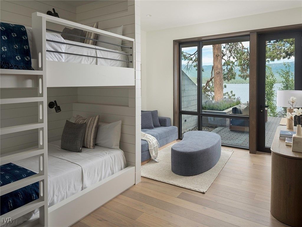 This interior shot showcases a modern bedroom with a built-in bunk bed featuring a white frame and clean lines. The room also includes a cozy seating area with a blue sofa and ottoman, complemented by a large window offering a view of a lake and outdoor patio. The hardwood flooring and neutral color palette create a serene and inviting atmosphere.