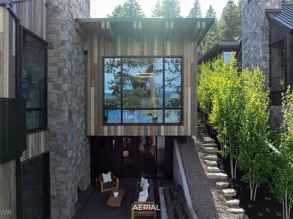 This is a striking front view of a modern home, showcasing a blend of stone and wood siding. A large window reflects the surrounding landscape, and an outdoor patio area with contemporary furniture adds to the appeal. Stone steps lead up the side of the house, framed by lush greenery.