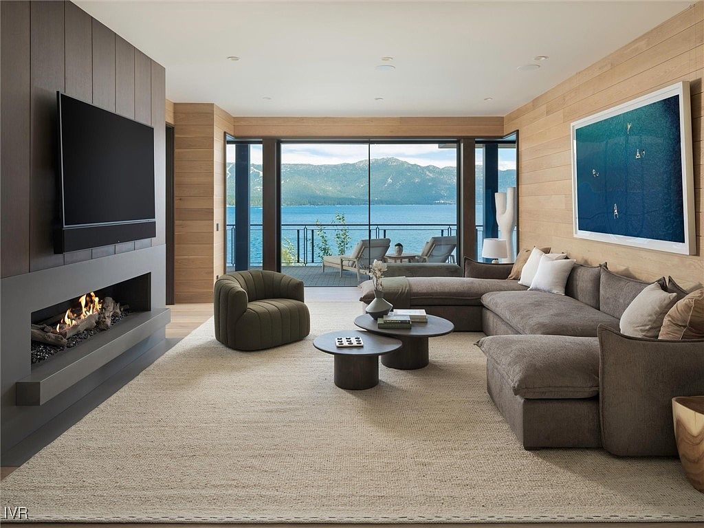 This is a beautifully designed living room featuring a modern fireplace with a large television above it, a comfortable sectional sofa, and a stylish round coffee table. Large windows offer stunning views of a lake and mountains, creating a serene and inviting atmosphere. The room is decorated in neutral tones with wood paneling, enhancing the natural light and creating a warm, luxurious feel.