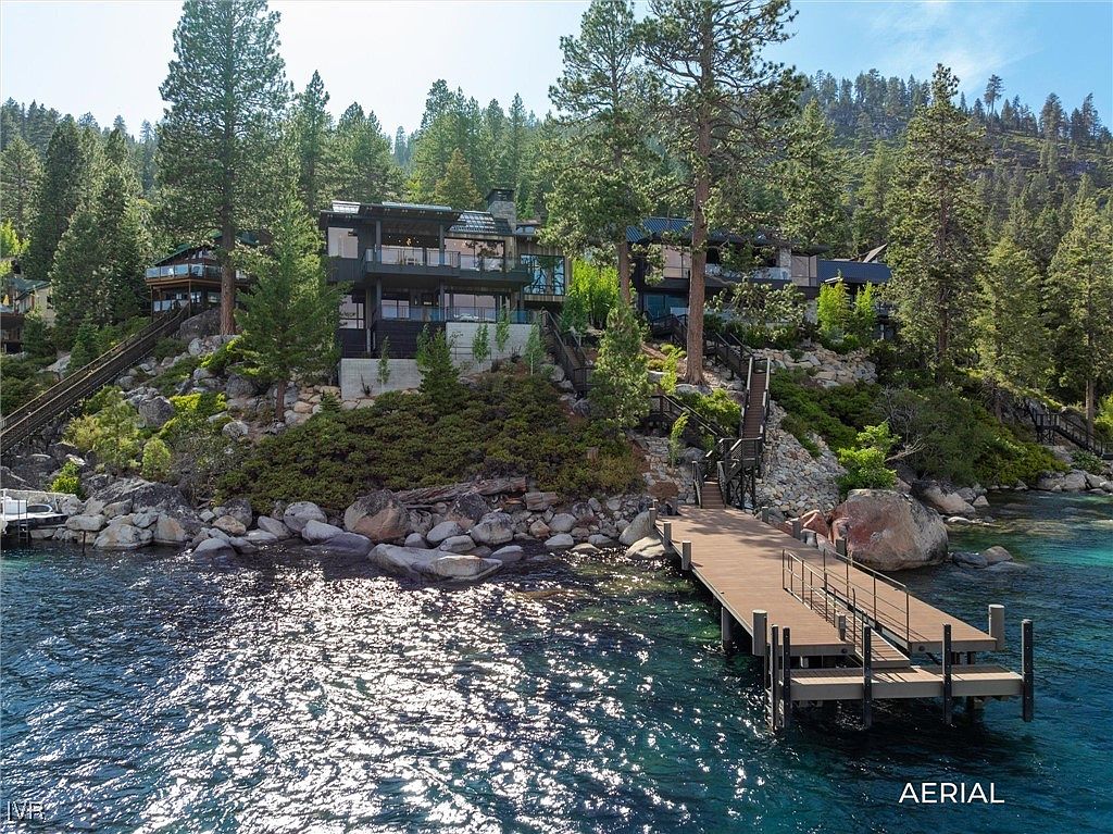 This aerial view showcases a luxurious waterfront property nestled among lush trees. The multi-level home features expansive windows and balconies, offering stunning views of the lake. A private dock extends into the clear, turquoise water, providing direct access for boating and other water activities.