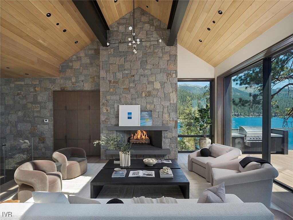 This is a spacious living room featuring a stone fireplace, wood-paneled ceiling, and large windows with a view of a lake and mountains. The room is furnished with comfortable seating, including armchairs and a sofa, arranged around a dark coffee table. The overall style is modern and luxurious, creating a cozy and inviting atmosphere.