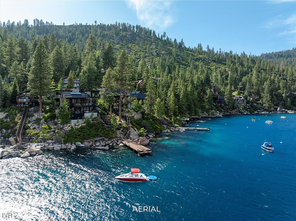 This aerial view showcases a stunning waterfront property nestled amidst lush greenery. The residence features modern architecture with expansive windows, seamlessly blending with the natural landscape. A private dock extends into the crystal-clear turquoise water, where a boat is anchored, creating an inviting scene for potential buyers seeking a luxurious lakeside retreat.