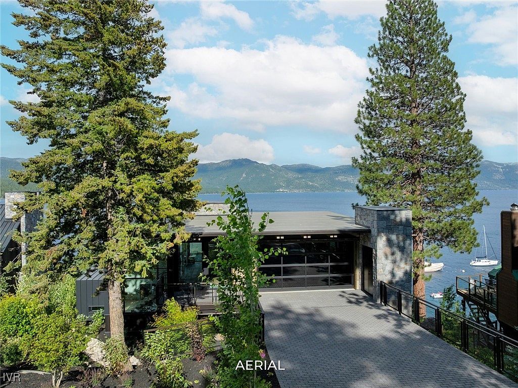 This aerial view showcases a modern home with a striking garage featuring glass doors, set against a backdrop of a serene lake and mountains. The property is framed by mature trees and lush landscaping, adding to its privacy and natural beauty. A paved driveway leads to the garage, enhancing the property's curb appeal.