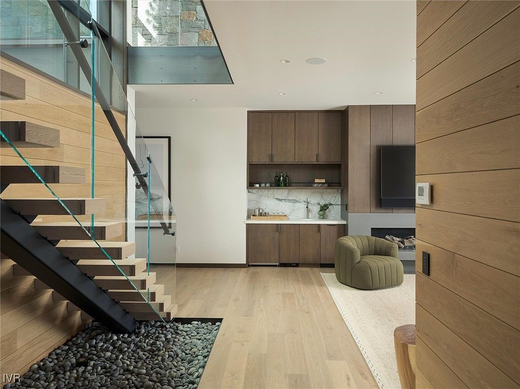 This interior shot showcases a modern living room with a unique staircase design featuring glass railings and a rock garden underneath. The room includes built-in cabinetry with a marble countertop, a fireplace, and a comfortable green armchair. The hardwood flooring and natural light create a warm and inviting atmosphere.