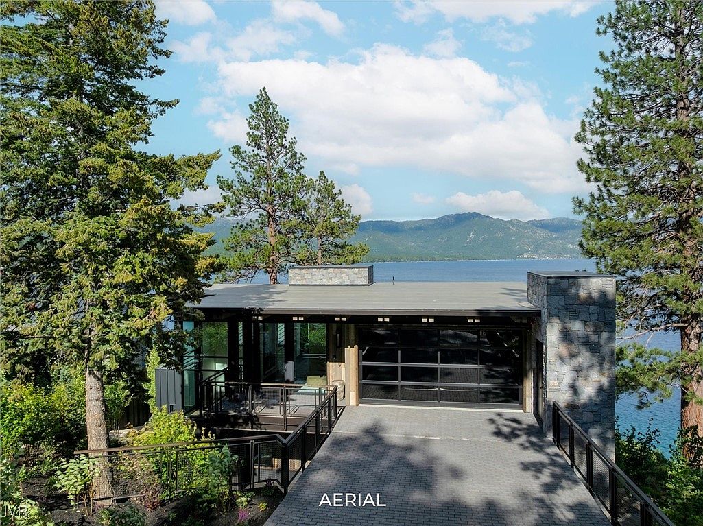 This aerial view showcases a modern home with a flat roof, stone accents, and a glass-paneled garage door. The property is nestled among mature trees and overlooks a serene lake with mountains in the background. A paved driveway leads to the garage, enhancing the property's curb appeal.