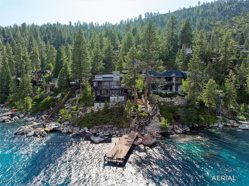 This aerial view showcases a luxurious waterfront property nestled among lush trees. The modern homes feature dark exteriors with large windows, offering stunning views of the lake. A private dock extends into the clear, turquoise water, creating an inviting scene for potential buyers seeking a serene and upscale lakeside retreat.