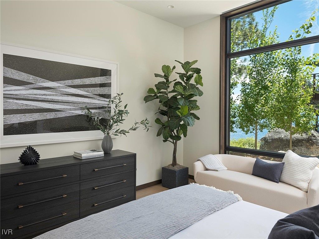 This is a well-lit primary bedroom featuring a dark wood dresser with modern art above it, a comfortable-looking daybed by a large window with a view of trees and water, and a glimpse of the bed with a gray throw. The room has a contemporary style with a focus on natural light and a calming color palette, creating a serene and inviting atmosphere.
