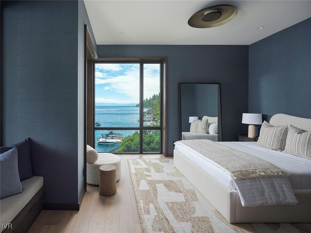 This is a primary bedroom featuring a large window with a view of the water, a king-sized bed with neutral linens, and a full-length mirror. The walls are a dark blue, and the flooring is light wood. The room has a modern and luxurious feel, with a focus on the view and natural light.