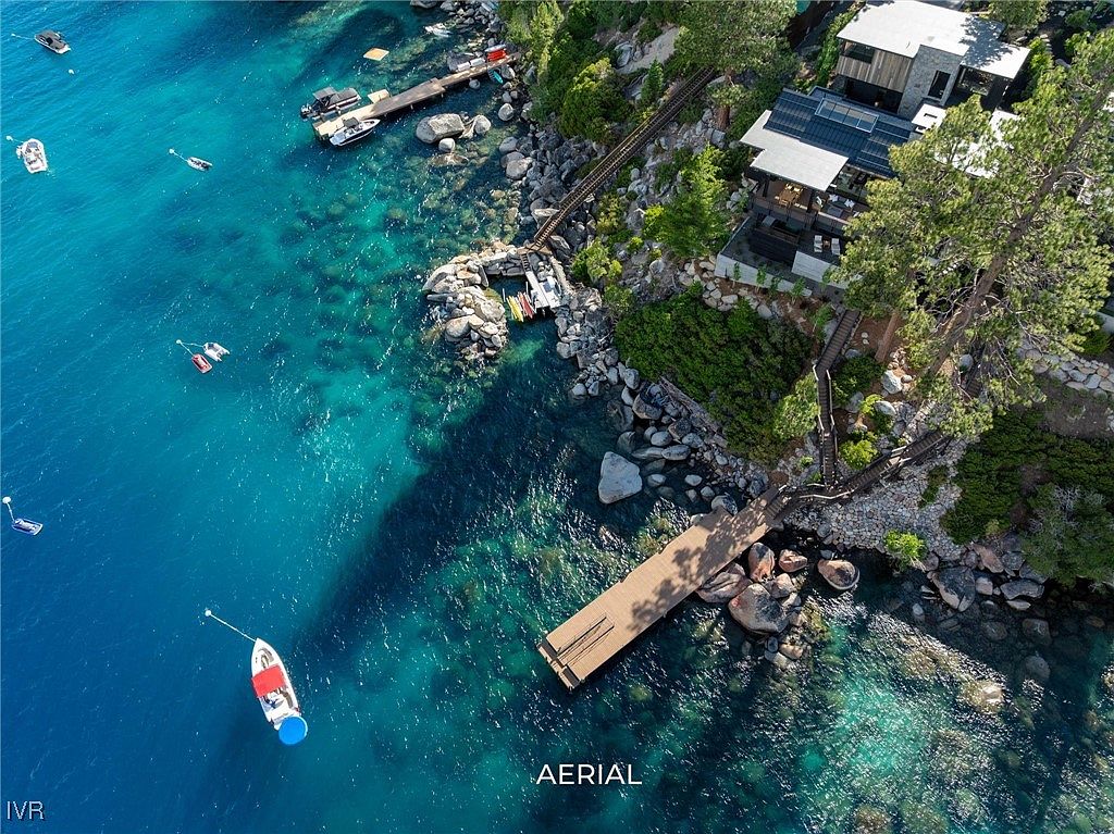 This aerial shot showcases a stunning waterfront property with a modern house nestled among trees and rocks. The property features multiple docks extending into the clear, turquoise water, with boats moored nearby. The landscape is a mix of natural rock formations and lush greenery, creating a serene and luxurious atmosphere.