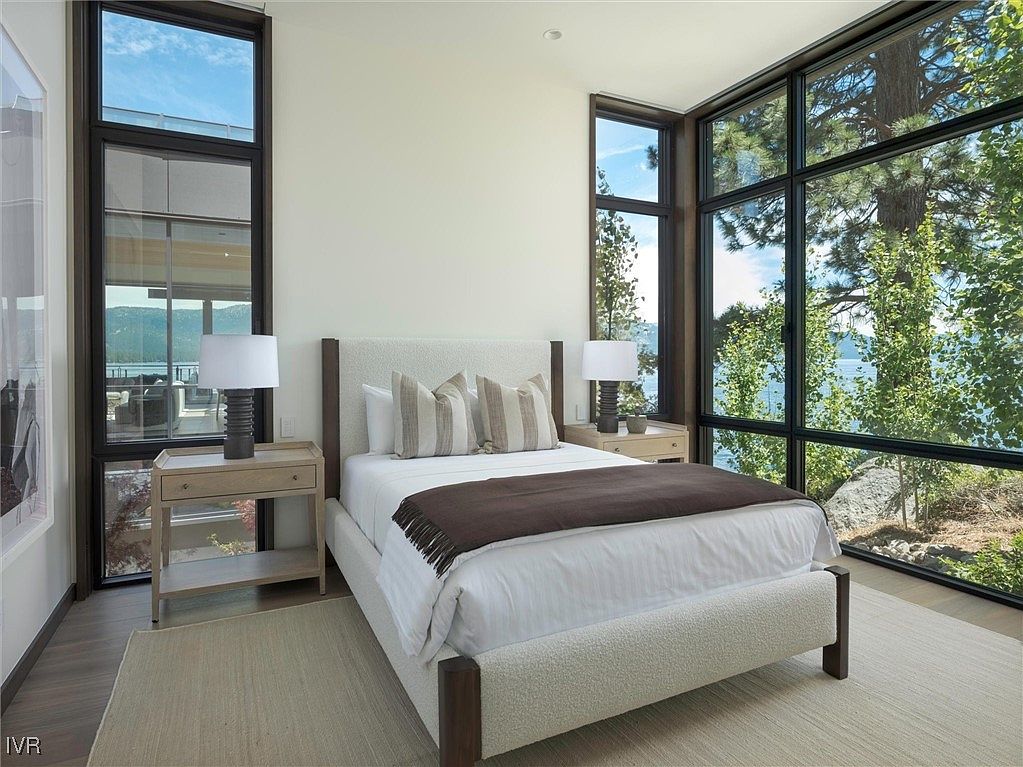 This is a primary bedroom featuring a large bed with a textured headboard and a brown throw blanket. The room has large windows offering a view of the lake and surrounding trees. Two nightstands with lamps flank the bed, and a neutral-colored rug covers the floor, creating a serene and inviting atmosphere.