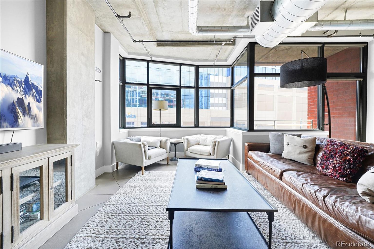 This is an interior shot of a modern living room. The room features a brown leather sofa, two white armchairs, and a black coffee table with books on top. Large windows offer city views, and the room is decorated with a patterned rug and a TV on a cabinet. The overall style is contemporary and industrial, with exposed concrete ceilings and ductwork.