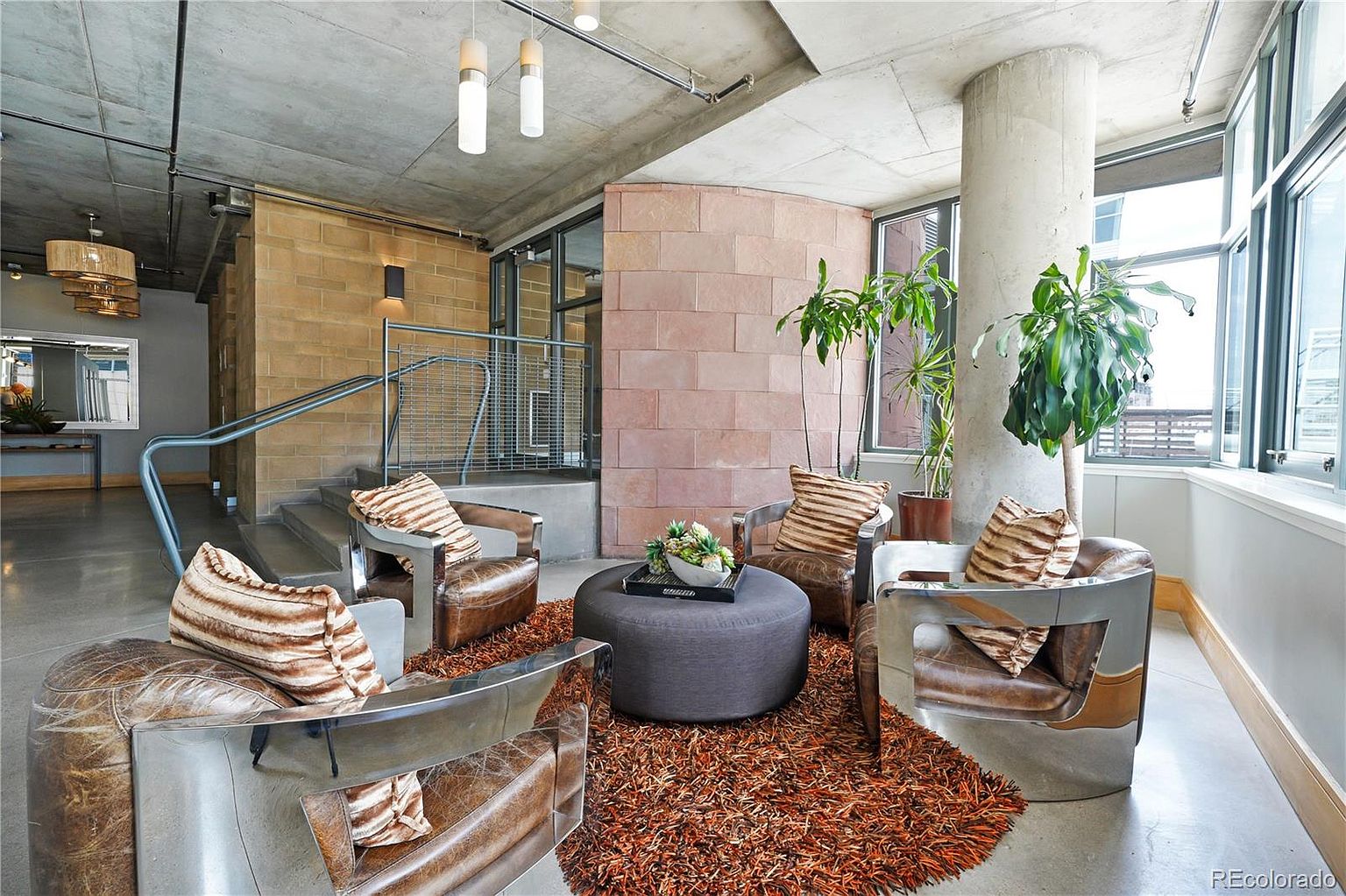 This interior shot showcases a stylish living room area with a modern industrial aesthetic. The space features four leather and metal armchairs arranged around a gray ottoman on a textured orange rug. Exposed concrete ceilings, brick accents, and large windows contribute to the room's unique character, creating a sophisticated and inviting atmosphere.