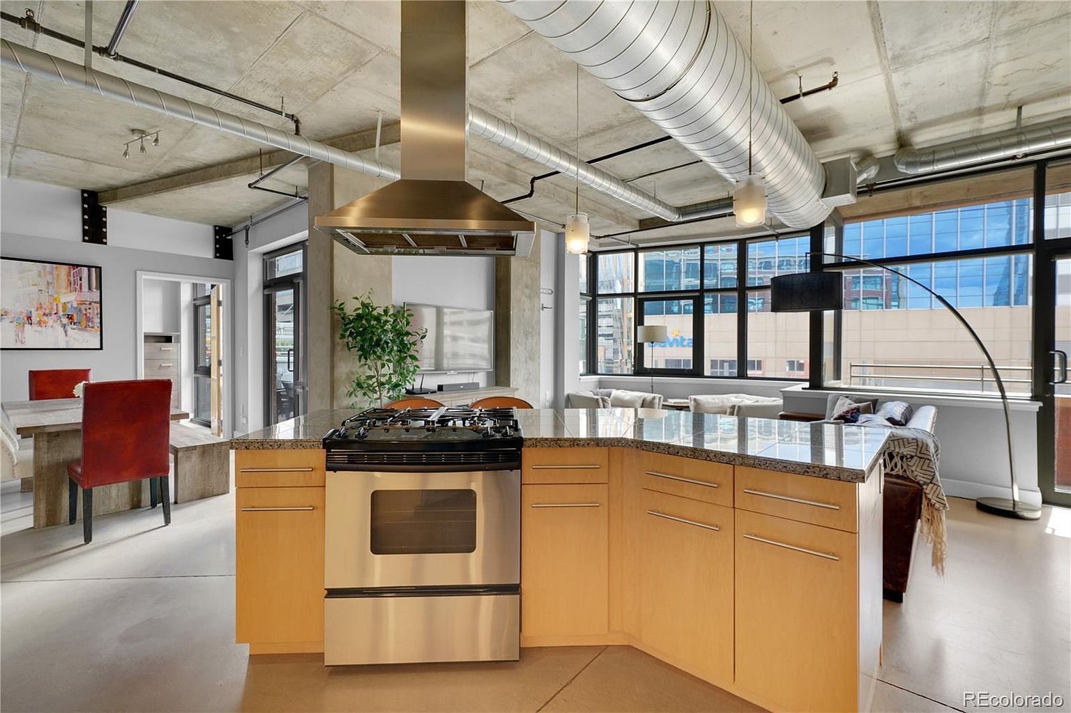 This interior shot showcases a modern kitchen with an island featuring a gas range and stainless steel oven. The kitchen boasts light wood cabinetry and granite countertops, complemented by stainless steel appliances and a unique exposed ductwork ceiling. Large windows provide ample natural light and views of the cityscape, blending the kitchen seamlessly into the adjacent living area.