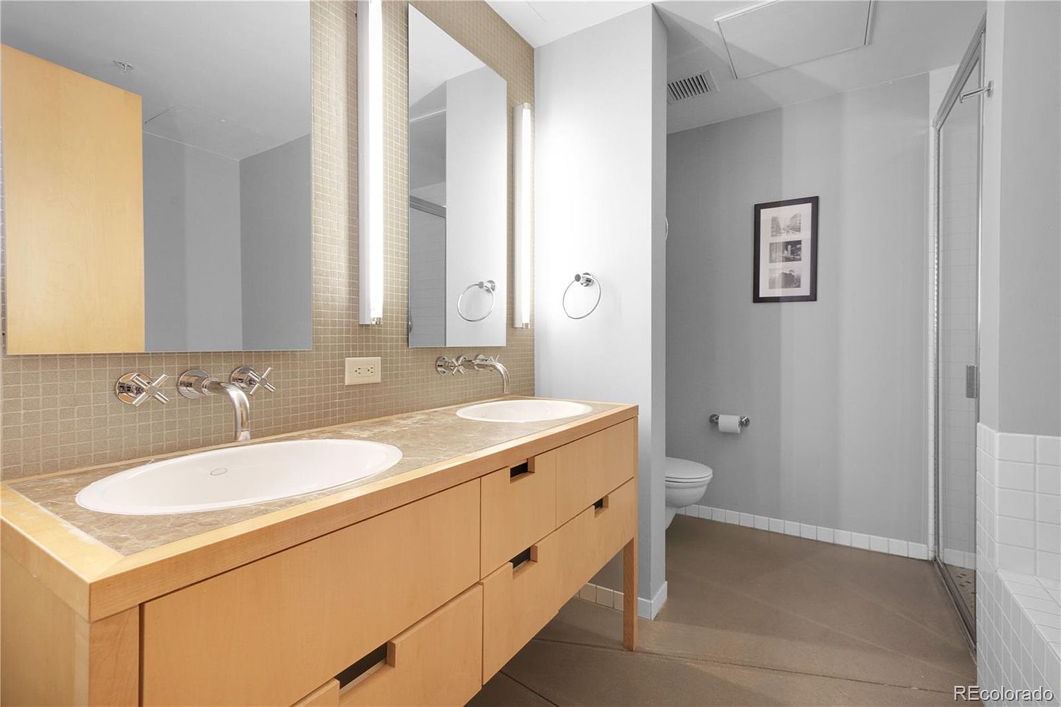 This is a well-lit primary bathroom featuring a double vanity with modern fixtures and a light wood finish. The countertop is a neutral stone, and the backsplash is tiled. A separate toilet and shower area are visible in the background, contributing to a spacious and functional design.