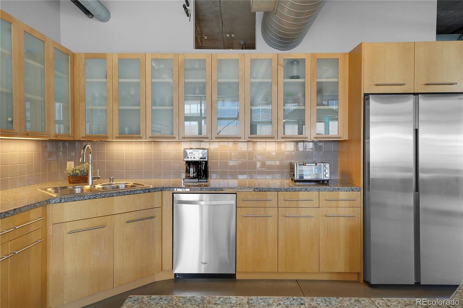 This is a well-lit kitchen featuring light wood cabinetry with frosted glass fronts on the upper cabinets. Stainless steel appliances, including a refrigerator, dishwasher, and toaster, add a modern touch. The countertops are granite, and the backsplash is a neutral tile, creating a clean and functional space.
