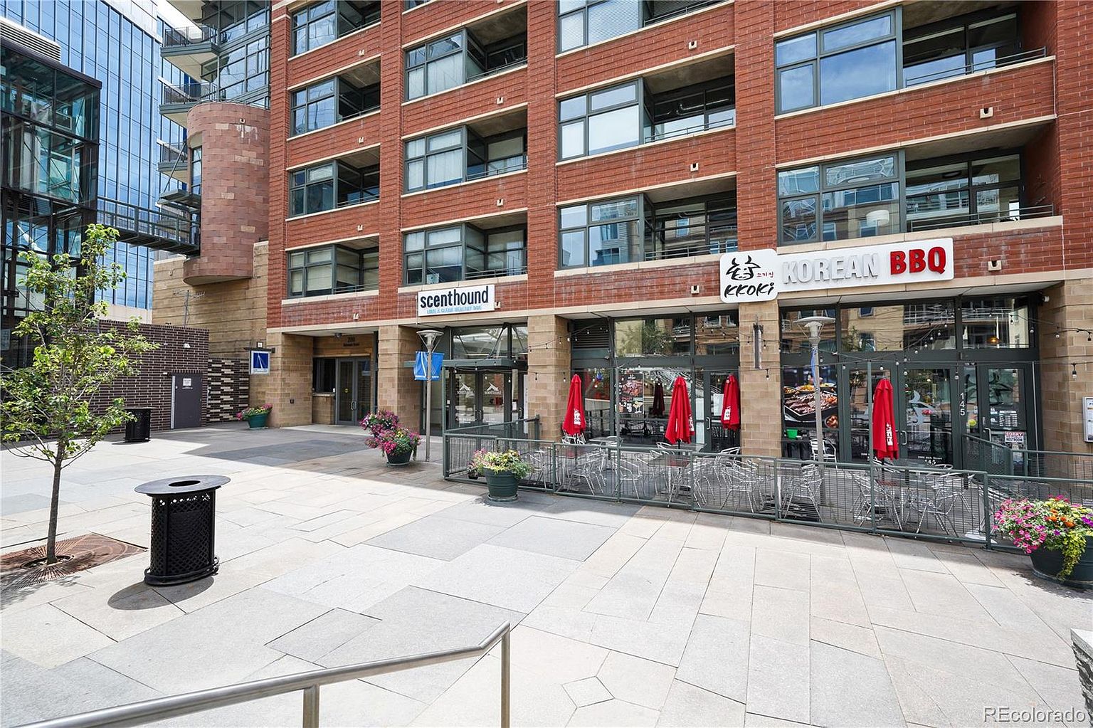 This is a front exterior view of a multi-story brick building with commercial spaces on the ground floor. The building features large windows, balconies, and signage for businesses like 'scenthound' and 'KOREAN BBQ'. The exterior space includes a paved walkway, outdoor seating with red umbrellas, and landscaping with flowers and trees.