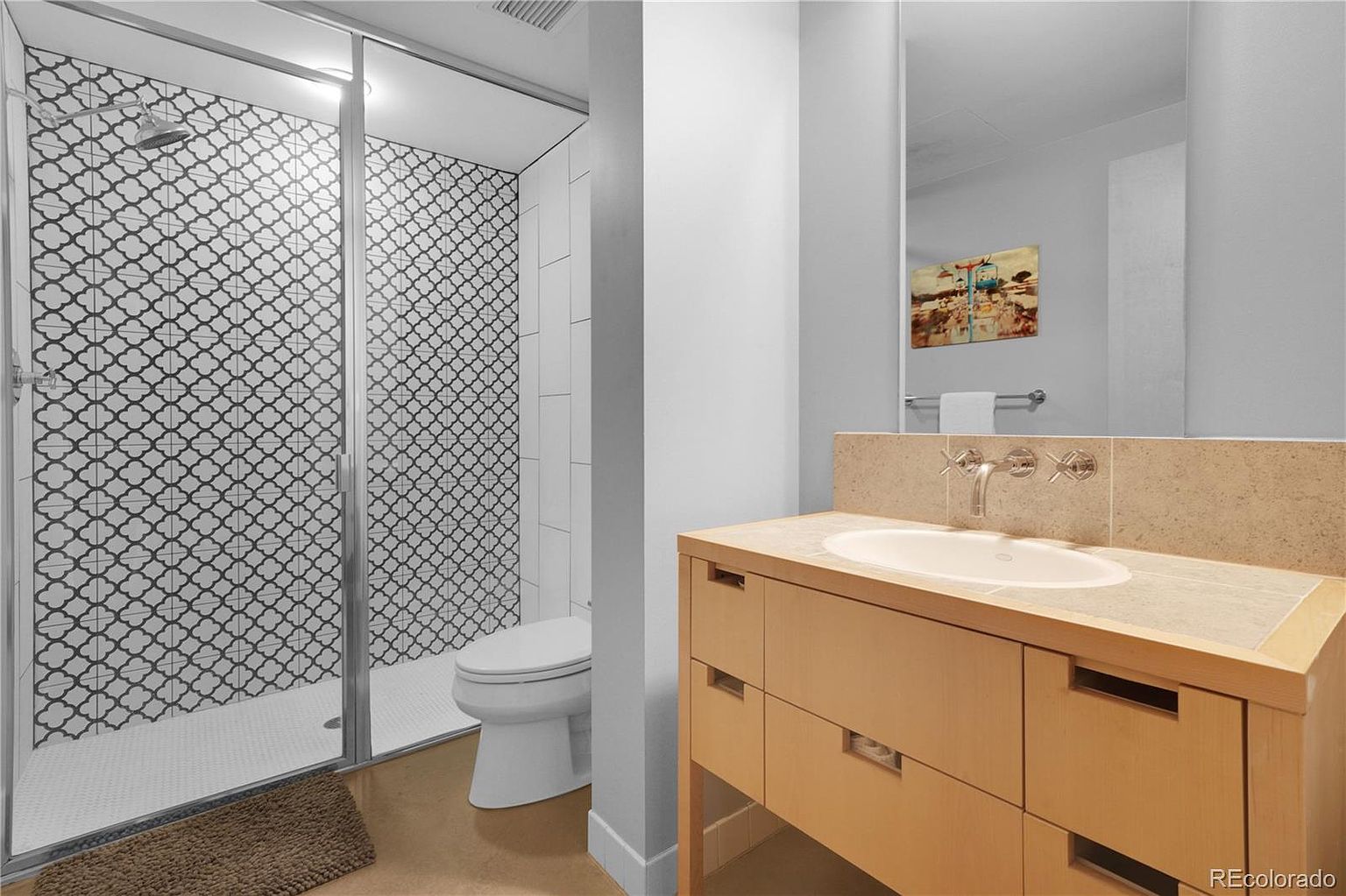 This is a well-lit bathroom featuring a modern design. The shower area is enclosed with glass and showcases patterned tiles, while the rest of the bathroom has a neutral color scheme. A wooden vanity with a light-colored countertop and a mirror above it complete the space, creating a clean and contemporary look.