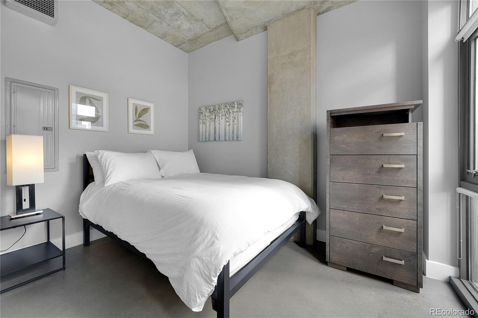 This is a well-lit primary bedroom featuring a bed with white linens, a dark-colored bed frame, and a matching nightstand with a lamp. A tall dresser stands against the wall, and artwork adorns the walls, creating a serene and modern atmosphere. The room has a minimalist design with concrete accents.