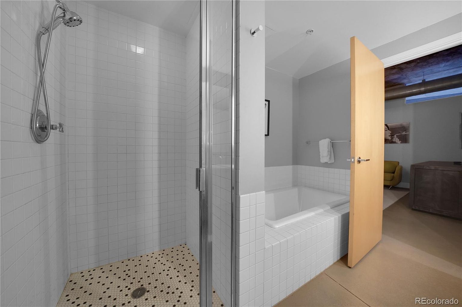 This is a well-lit primary bathroom featuring a walk-in shower with white tiled walls and a unique patterned floor. Adjacent to the shower is a bathtub encased in similar white tiles, with a light wood door partially open, leading to the rest of the primary suite. The overall aesthetic is clean and modern.