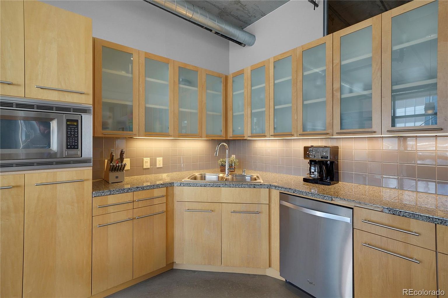 This is a well-lit kitchen featuring light wood cabinetry with frosted glass upper cabinets and stainless steel hardware. The countertops are granite, and the backsplash is tiled. Stainless steel appliances, including a microwave and dishwasher, are visible, contributing to a modern and functional space.