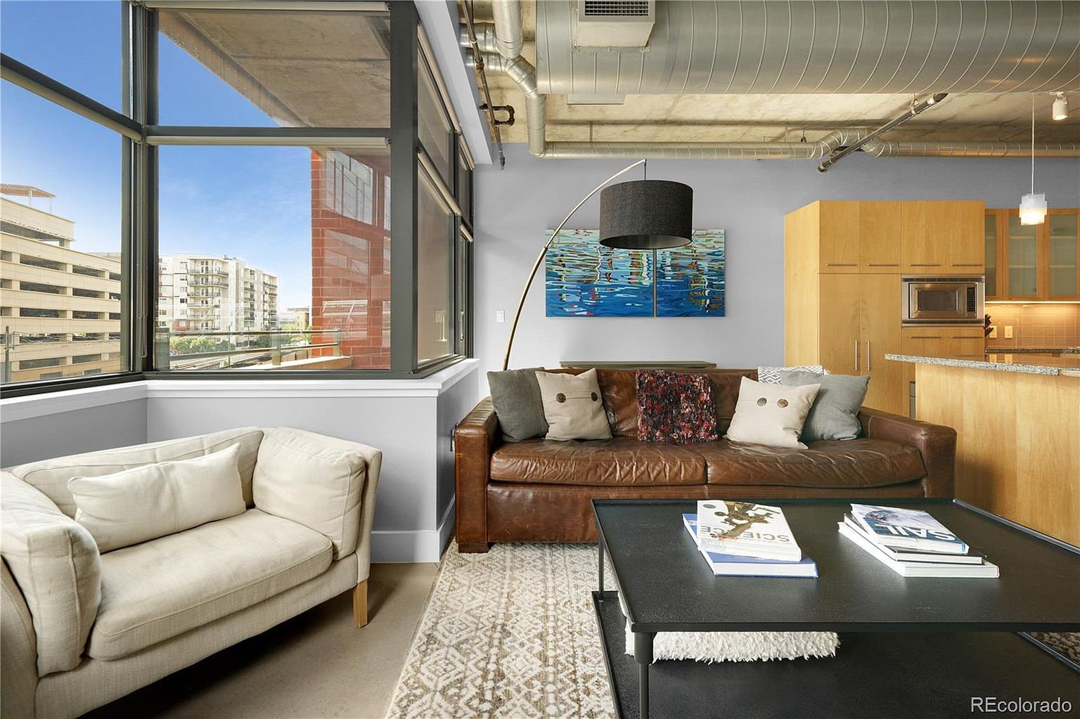This interior shot showcases a stylish living room with a blend of modern and industrial elements. A large window provides ample natural light and a view of the cityscape. The room features a comfortable leather sofa, a neutral-toned armchair, and a dark coffee table, complemented by a patterned area rug, creating a cozy and inviting atmosphere.