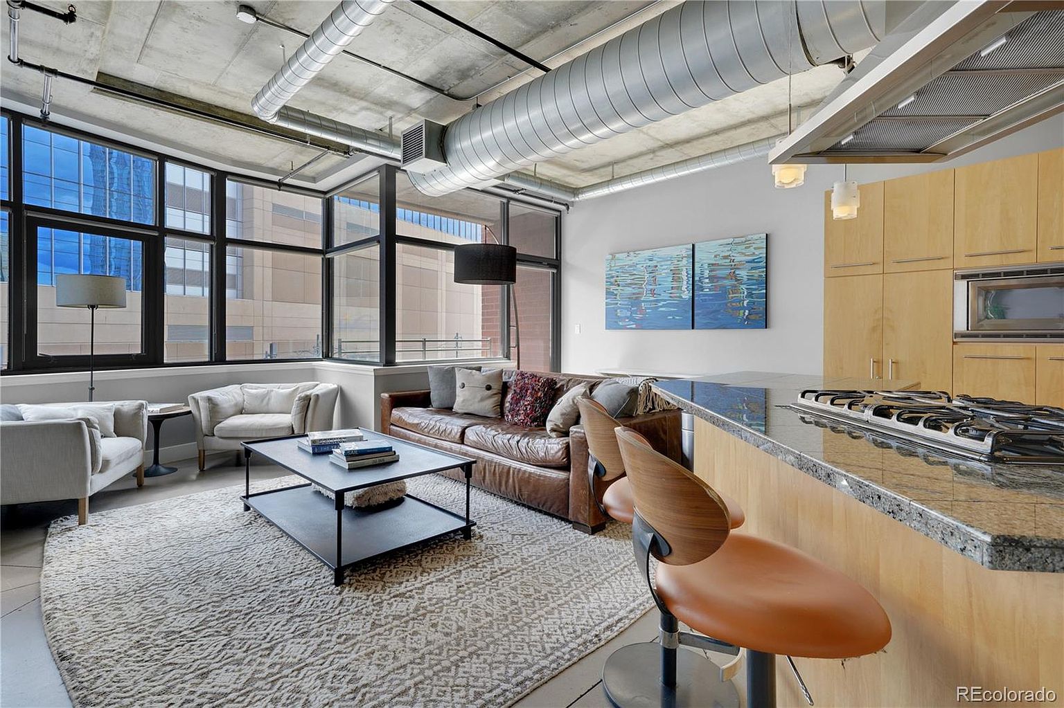 This is an interior shot of a modern living room and kitchen area. The living room features a leather sofa, two armchairs, a coffee table, and large windows offering city views. The kitchen area includes light wood cabinetry, granite countertops, and stainless steel appliances, creating a stylish and functional living space.