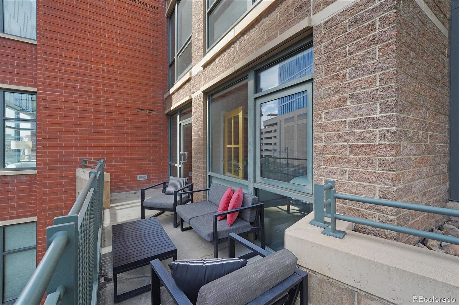 This image showcases a stylish balcony or patio area, furnished with modern outdoor seating and a coffee table. The space is enclosed by a metal railing and features a mix of brick and stone exterior walls, creating an urban yet inviting atmosphere. Large windows reflect the surrounding cityscape, adding depth and visual interest.