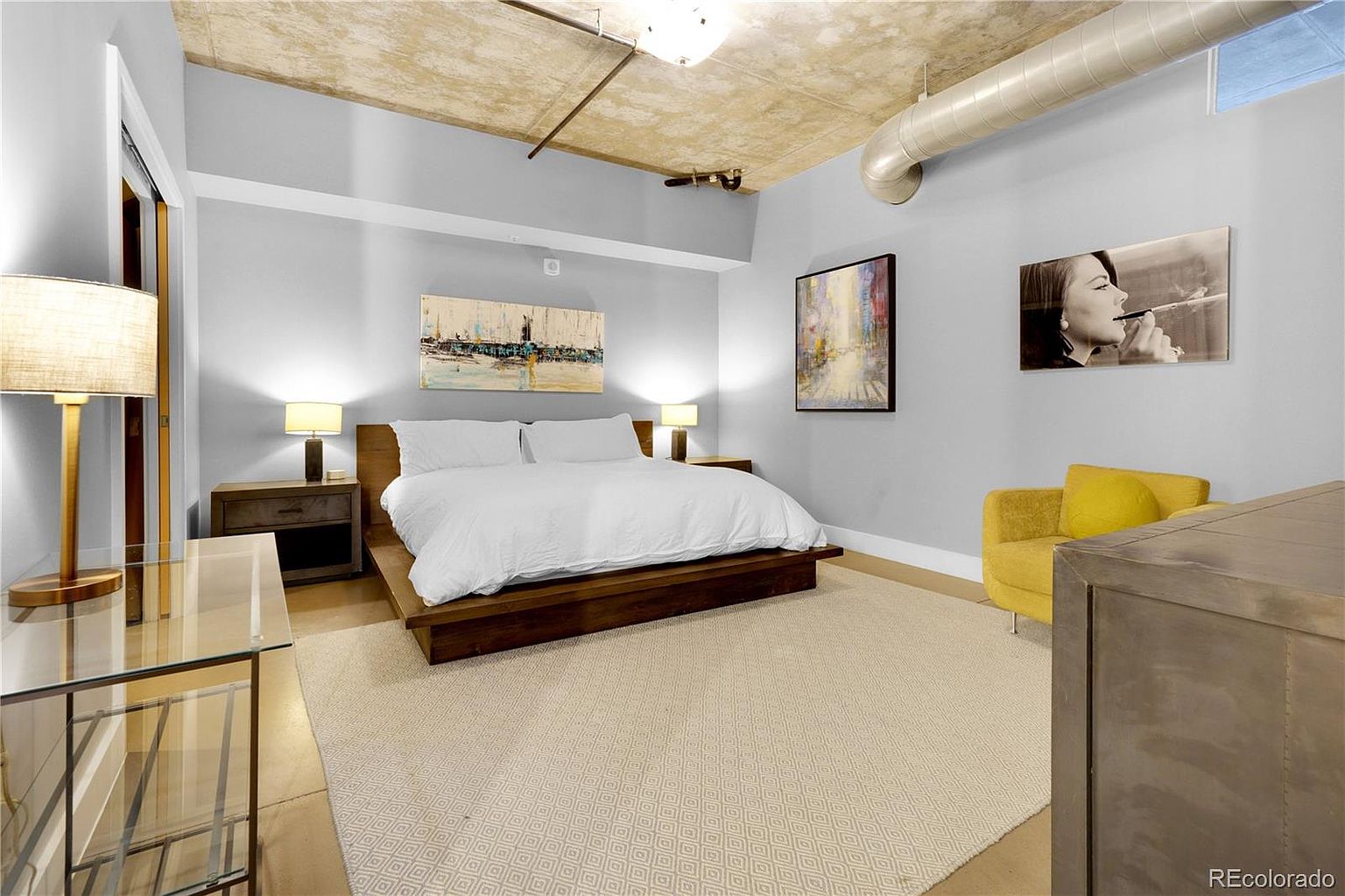 This is a primary bedroom featuring a large bed with white linens, flanked by wooden nightstands and lamps. Artwork adorns the walls, and a yellow armchair adds a pop of color. The room has a modern, industrial feel with exposed ceiling elements and a neutral color palette, creating a serene and stylish atmosphere.