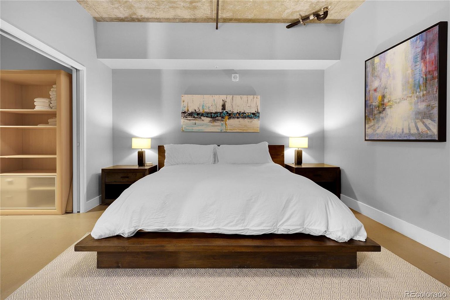 This is a well-lit primary bedroom featuring a large bed with white linens on a dark wood platform. Two nightstands with lamps flank the bed, and artwork hangs above the bed and on the adjacent wall. A closet is visible to the left, and the room has a modern, minimalist aesthetic.