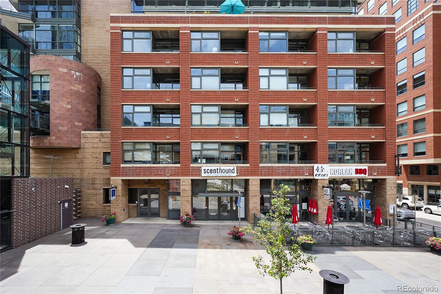 The image showcases the front exterior of a brick building, likely a condominium or apartment complex, featuring multiple stories with large windows and balconies. The ground level houses commercial spaces, including a restaurant with outdoor seating. The building's facade is well-maintained, and the surrounding plaza is clean and inviting, suggesting a desirable urban living environment.