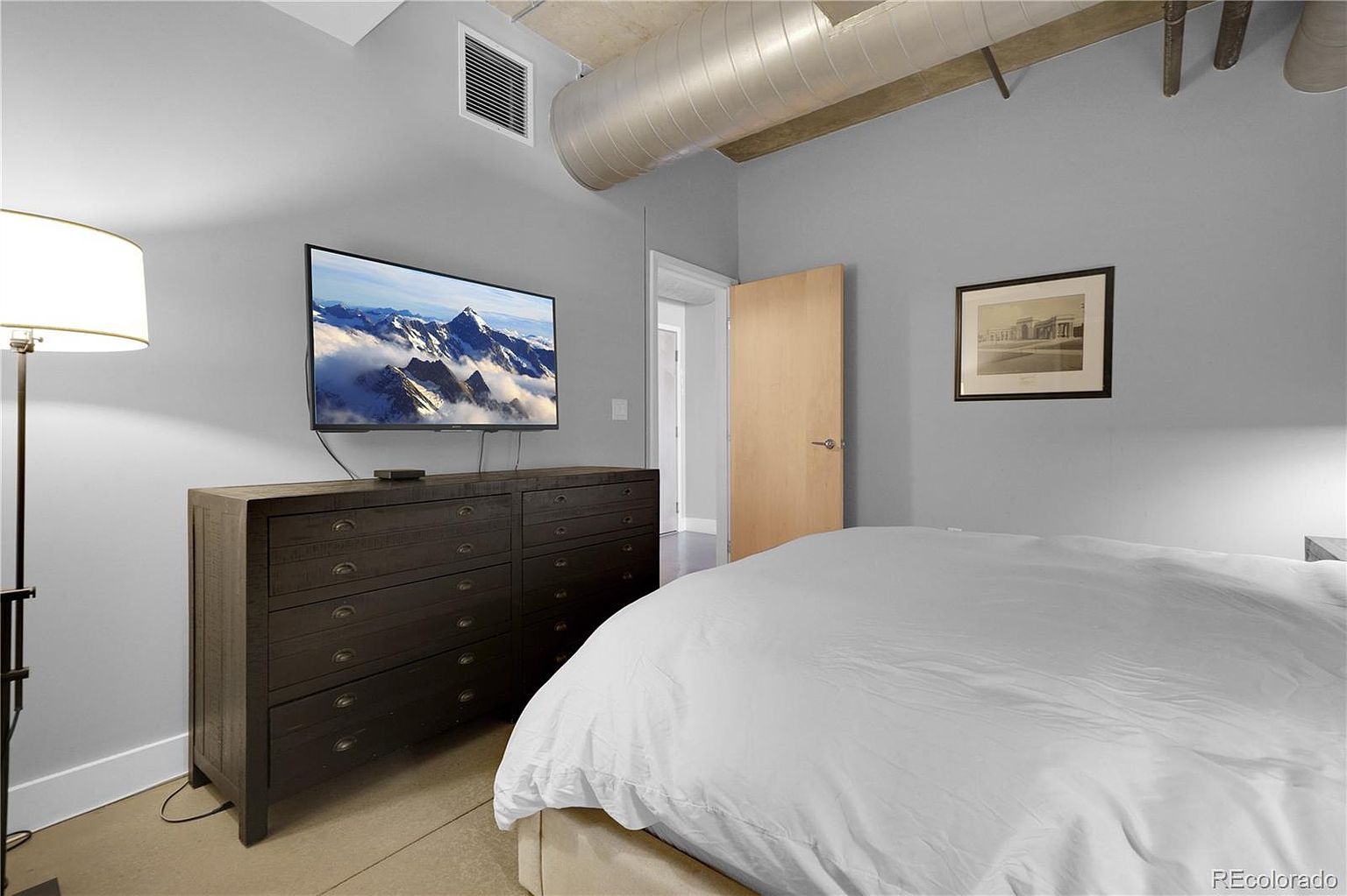 This is a primary bedroom featuring a large bed with white linens, a dark wood dresser with a flat-screen TV mounted above it, and a framed picture on the wall. The room has a modern, minimalist style with gray walls and exposed ductwork on the ceiling. The perspective is from the foot of the bed, looking towards the dresser and TV.