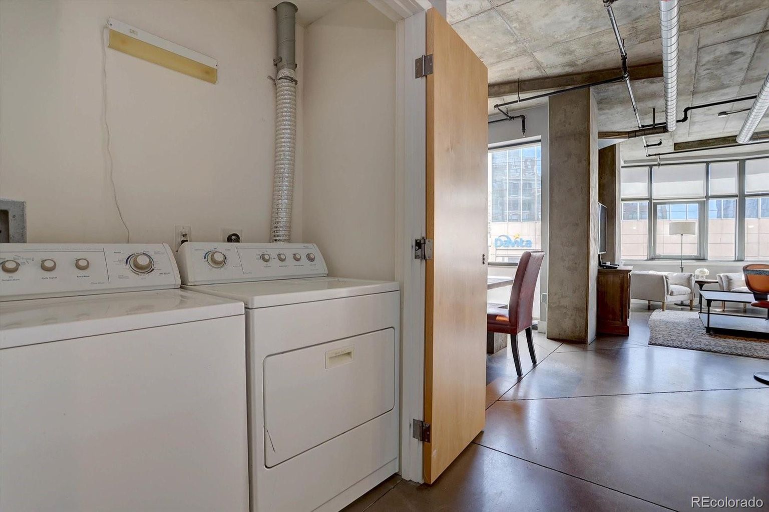 This image showcases a laundry room with a side-by-side washer and dryer set. The room is adjacent to a living space, visible through an open doorway, which features concrete ceilings and modern furniture. The laundry area is functional and well-lit, while the living space offers a glimpse of contemporary urban living.
