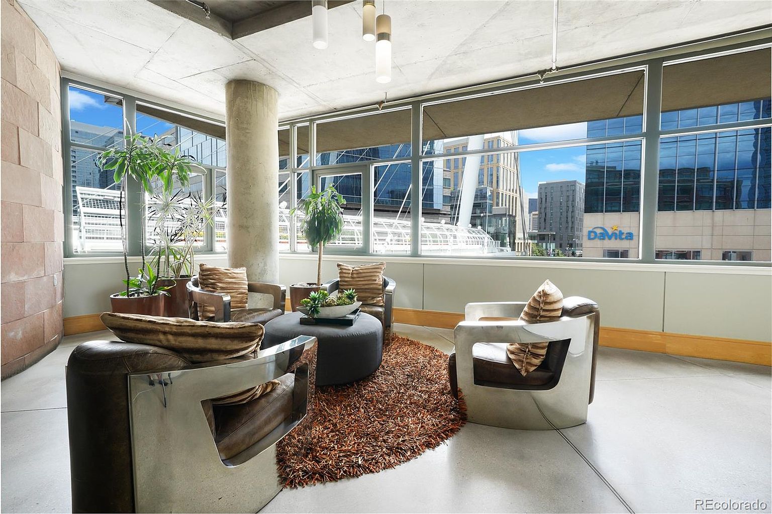 This interior shot showcases a modern living room with an industrial touch, featuring exposed concrete ceilings and a pillar. The room is furnished with unique, contemporary chairs, a round ottoman, and a textured brown rug, creating a cozy yet stylish atmosphere. Large windows offer a view of the cityscape, enhancing the urban living experience.