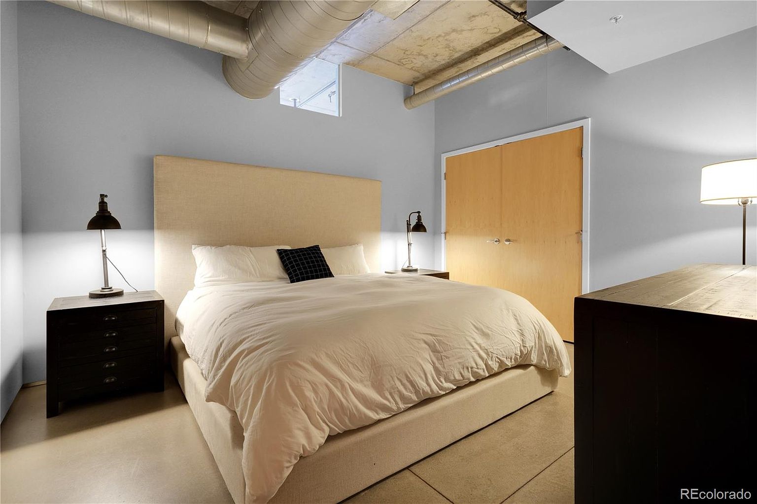 This is a primary bedroom featuring a large bed with a beige headboard and white bedding. The room has a modern, industrial feel with exposed ductwork on the ceiling and a minimalist design. There are bedside tables with lamps on either side of the bed, and a dark wood dresser is visible in the corner.