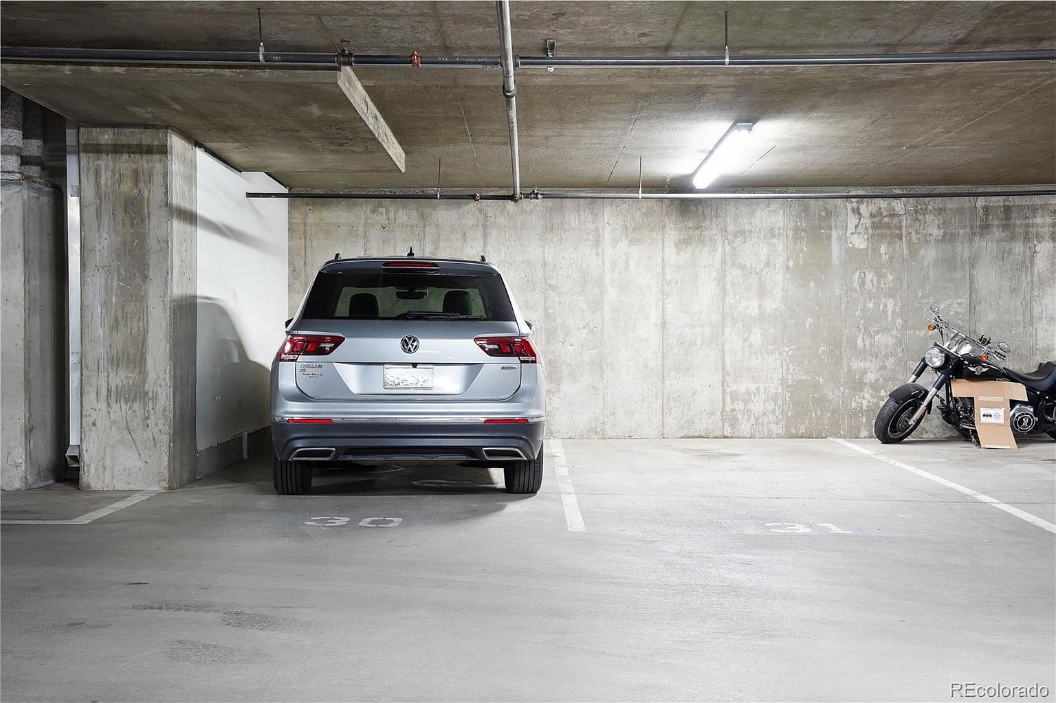 The image shows the rear view of a silver Volkswagen Tiguan parked in a concrete parking garage. The garage has exposed concrete walls and ceiling with visible pipes and lighting fixtures. A motorcycle is parked in the adjacent space, and parking space numbers are painted on the floor, creating a functional and utilitarian atmosphere.