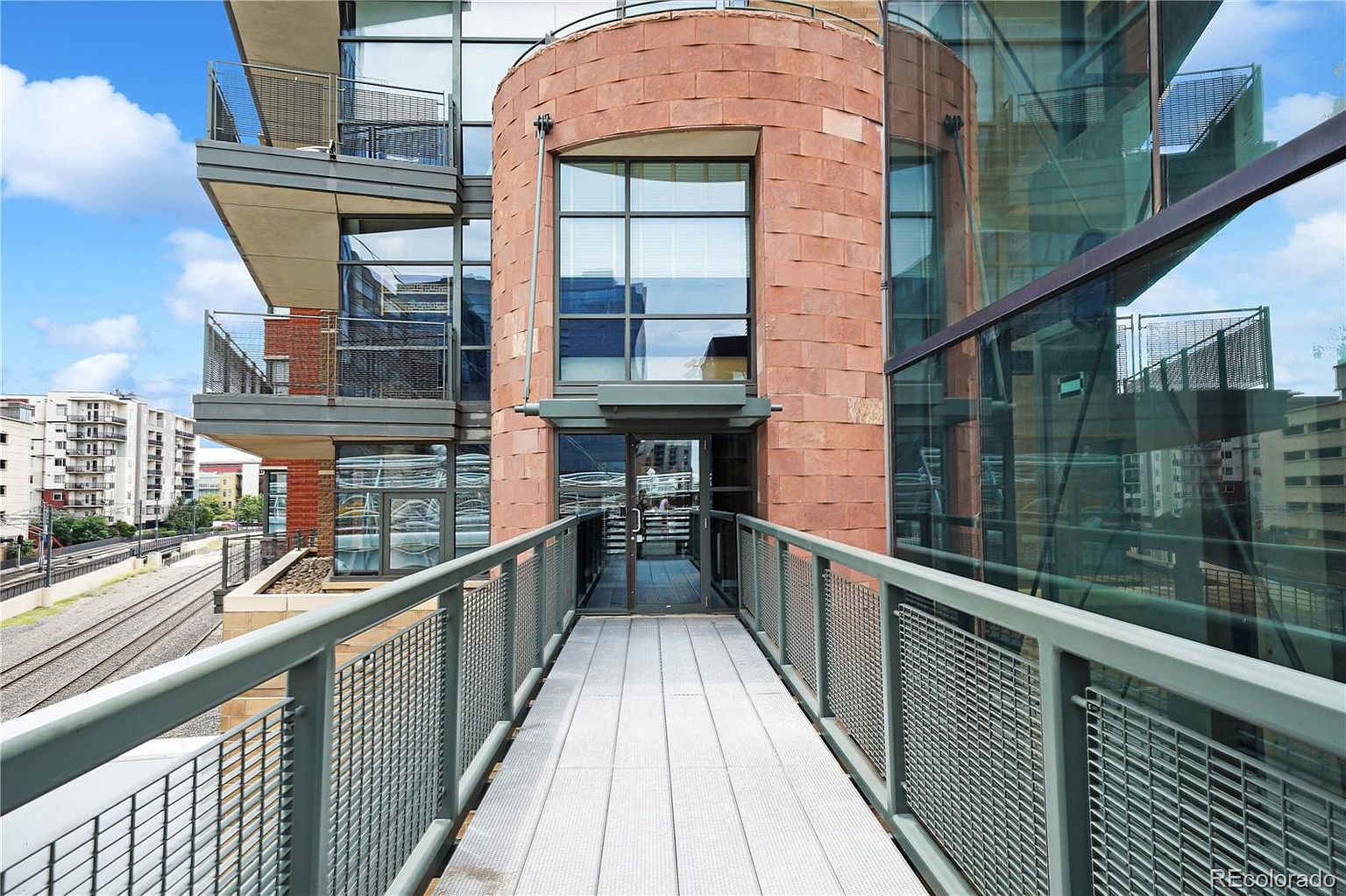 This image showcases a unique entryway to a modern building, featuring a pedestrian bridge leading to a glass-door entrance. The building's facade combines red brick and glass elements, creating a contemporary architectural style. The perspective is from the bridge, emphasizing the approach to the building and the surrounding urban environment.