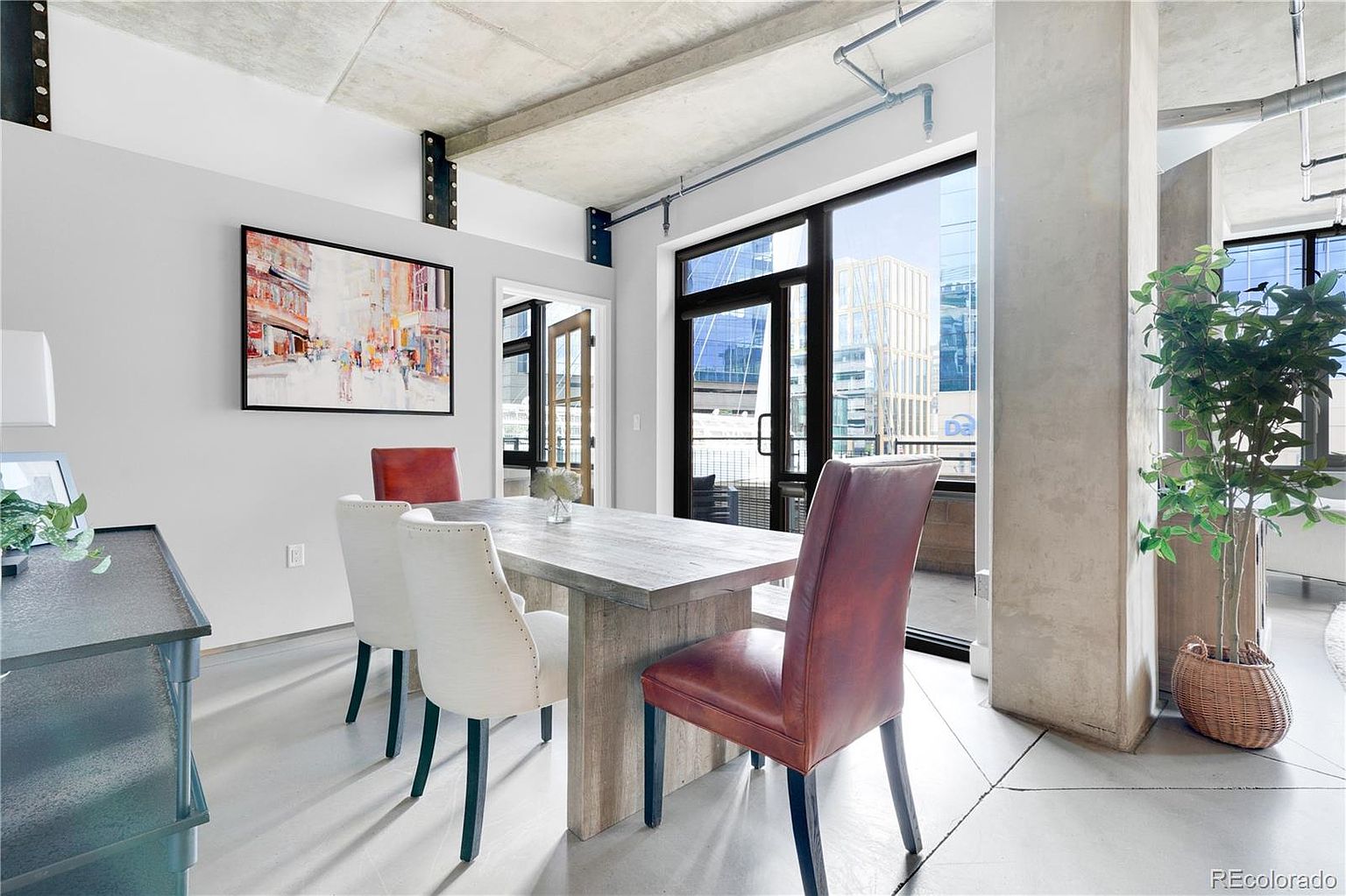 This interior shot showcases a modern dining room with a rustic-chic aesthetic. A large wooden dining table is surrounded by upholstered chairs, and a contemporary art piece hangs on the wall. Natural light floods the space through a large window, highlighting the concrete ceiling and industrial-style support beams, creating an open and airy atmosphere.