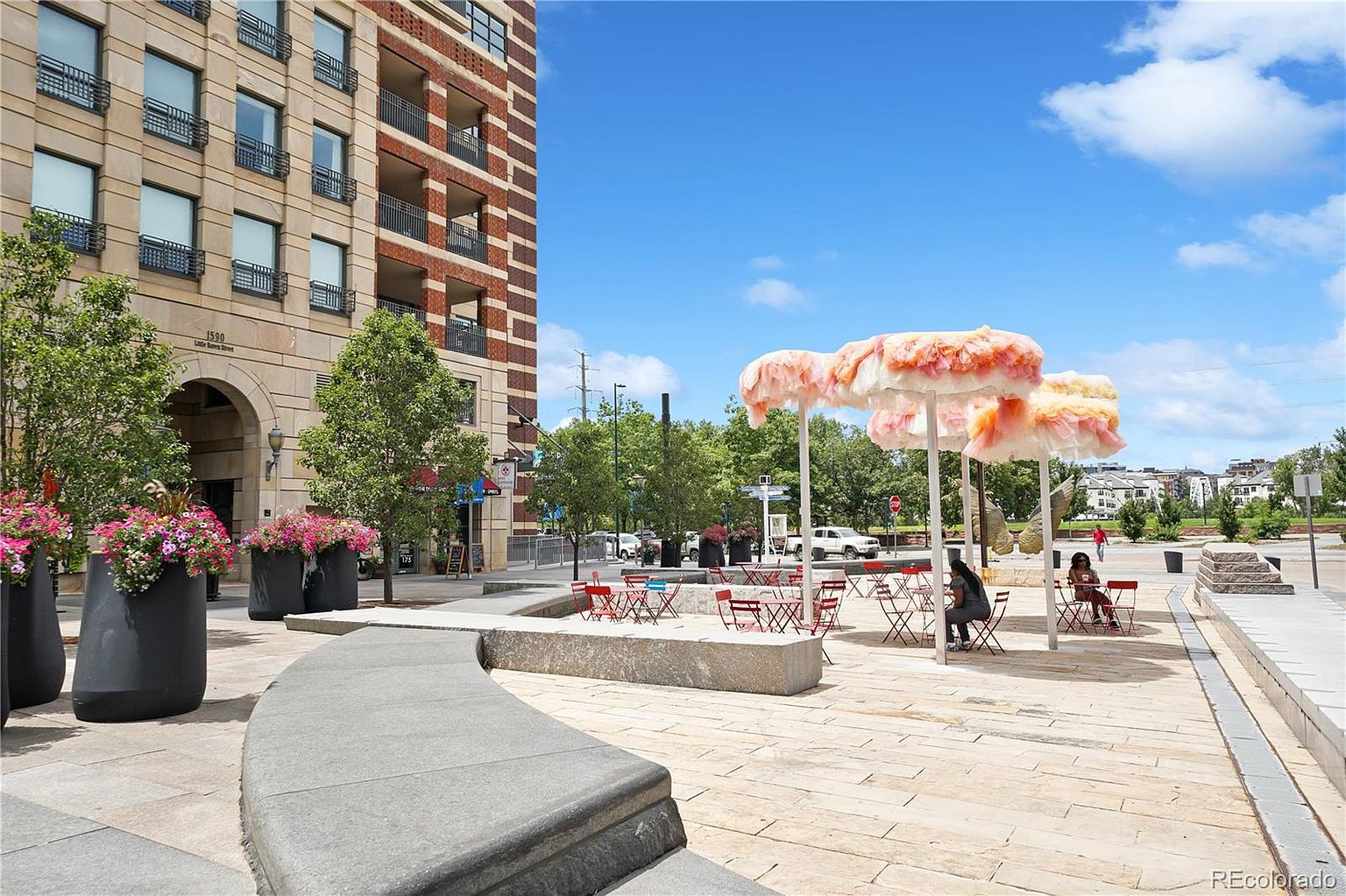 This image showcases a vibrant outdoor community space with modern art installations, seating areas, and lush greenery. The area features a mix of hardscaped surfaces and planters, creating an inviting atmosphere for residents and visitors. A tall building is visible in the background, suggesting an urban setting.