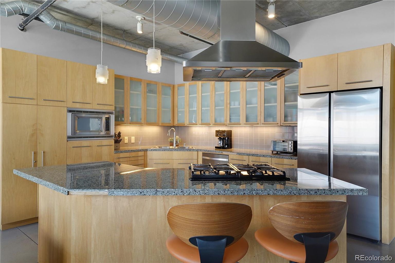 This is a modern kitchen featuring light wood cabinetry, stainless steel appliances, and a granite countertop island with a gas cooktop. The kitchen has a contemporary design with exposed ductwork and pendant lighting, creating an industrial-chic aesthetic. The perspective is from a medium shot, focusing on the island and the surrounding cabinetry.