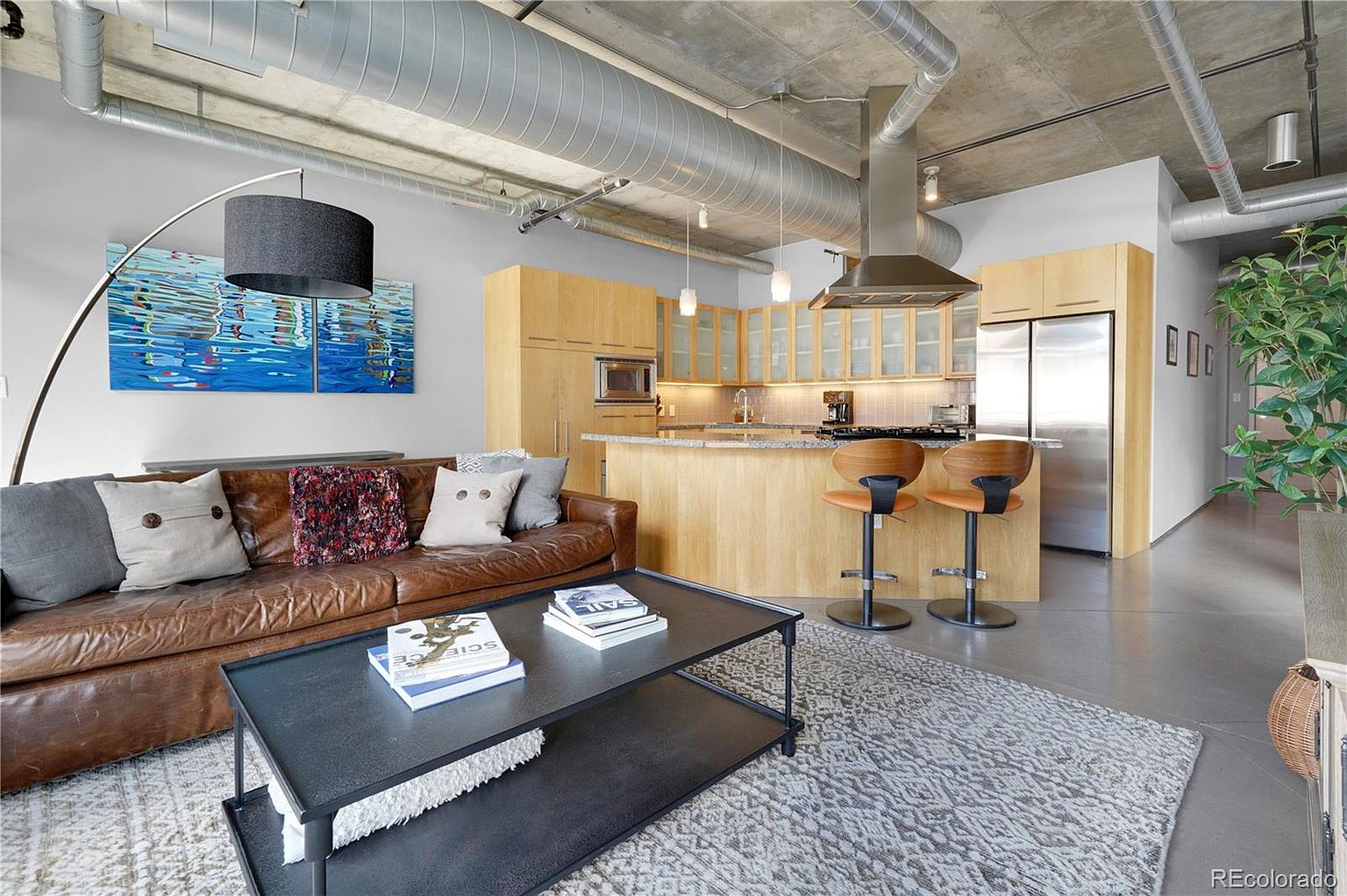 This is an interior shot of a modern living room and kitchen area. The living room features a brown leather sofa with decorative pillows, a black coffee table with books, and a gray patterned rug. The kitchen has light wood cabinetry, stainless steel appliances, and a breakfast bar with two stools. Exposed ductwork and a concrete ceiling give the space an industrial-chic feel.