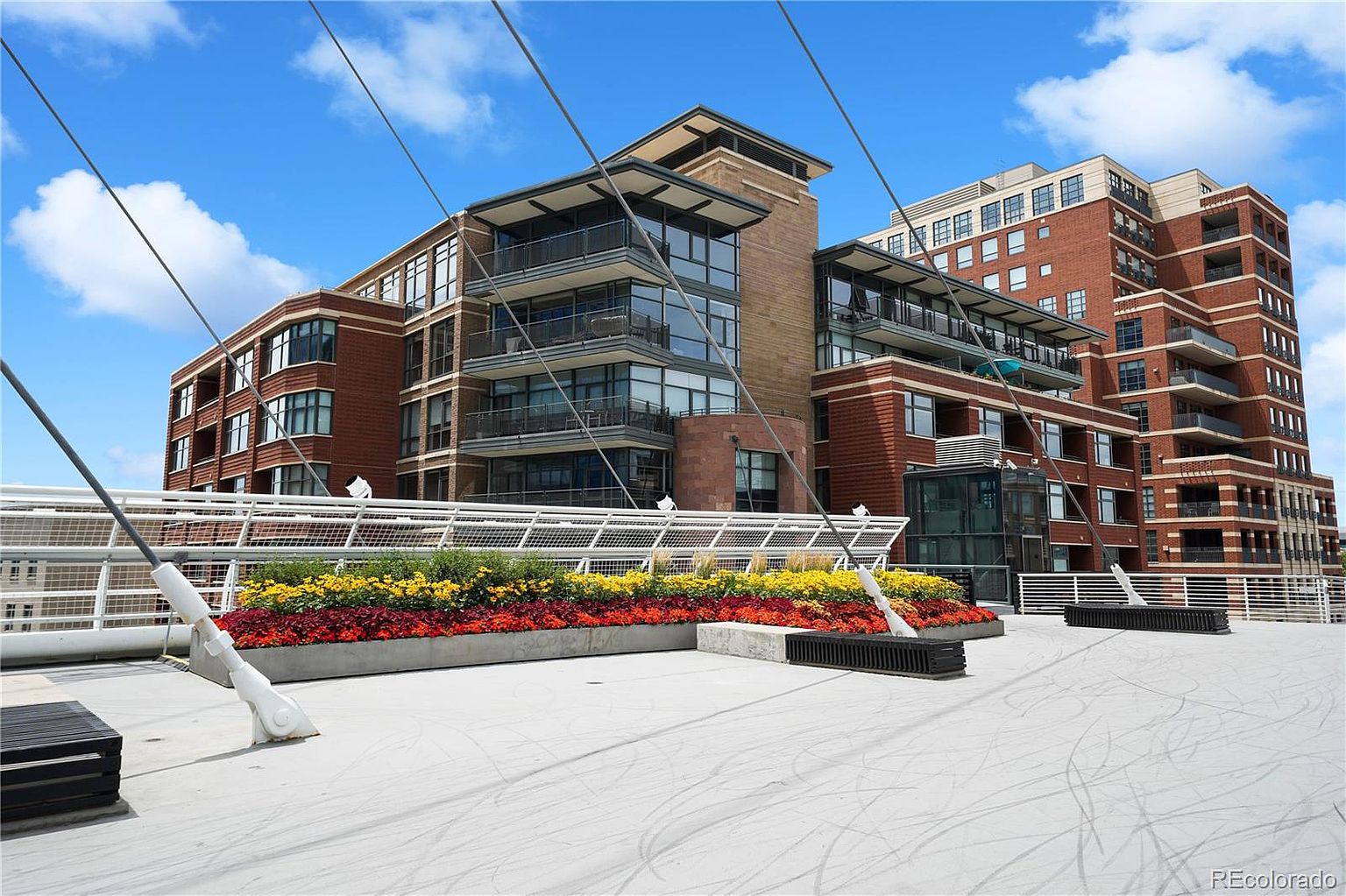 This image showcases a spacious patio or deck area of a modern residential building. The area features colorful flower beds, contemporary benches, and a unique architectural design with supporting cables. The backdrop includes a multi-story brick building with balconies, suggesting a high-end urban living environment.